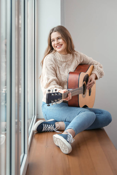Νεαρή γυναίκα με κιθάρα κάθεται στο περβάζι του παραθύρου στο σπίτι - Φωτογραφία, εικόνα