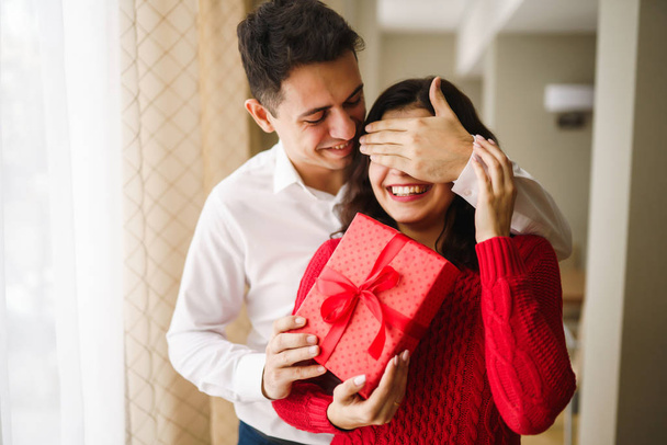 A young man gives his beloved a present and closes her eyes with a hand. Beautiful woman receivs a surprise gift box from her boyfriend at restaurant. Valentine's Day. Women's Day. Birthday concept. - Foto, Imagen