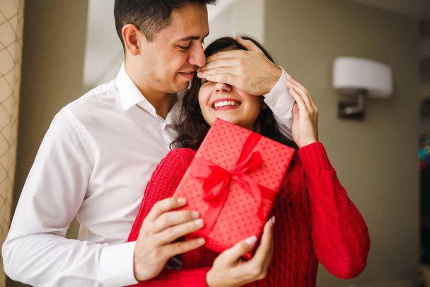 A young man gives his beloved a present and closes her eyes with a hand. Beautiful woman receivs a surprise gift box from her boyfriend at restaurant. Valentine's Day. Women's Day. Birthday concept. - Photo, image