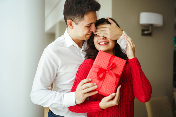 A young man gives his beloved a present and closes her eyes with a hand. Beautiful woman receivs a surprise gift box from her boyfriend at restaurant. Valentine's Day. Women's Day. Birthday concept. - Fotó, kép