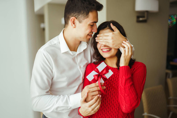 A young man gives his beloved a present and closes her eyes with a hand. Beautiful woman receivs a surprise gift box from her boyfriend at restaurant. Valentine's Day. Women's Day. Birthday concept. - Photo, Image