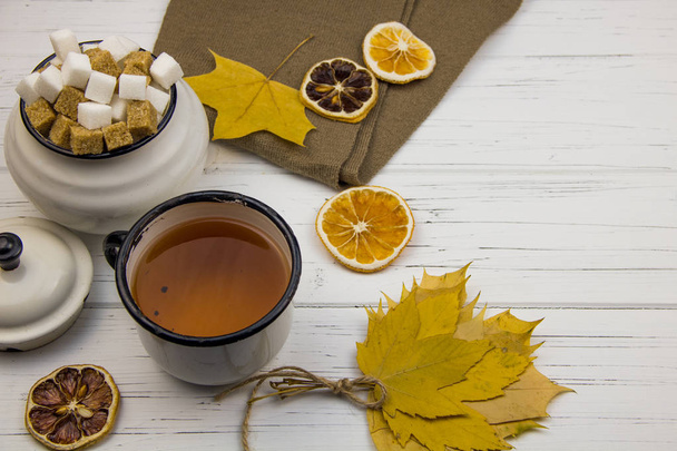 chá em uma xícara de alumínio e açúcar em cubos em uma tigela de açúcar de alumínio, cachecol marrom, folhas de bordo, laranjas secas e limão seco em um fundo de madeira leve
 - Foto, Imagem