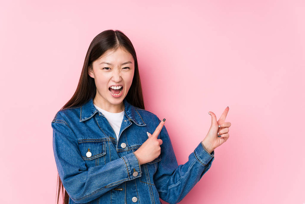 Молодая китаянка позирует на розовом фоне изолированно указывая указательными пальцами на пространство для копирования, выражая восхищение и желание
. - Фото, изображение