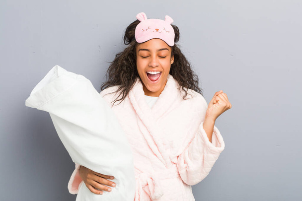 Νεαρή Αφροαμερικάνα που φοράει πιτζάμες και μάσκα ύπνου κρατώντας ένα μαξιλάρι ζητωκραυγάζει ανέμελη και ενθουσιασμένη. Έννοια νίκης. - Φωτογραφία, εικόνα