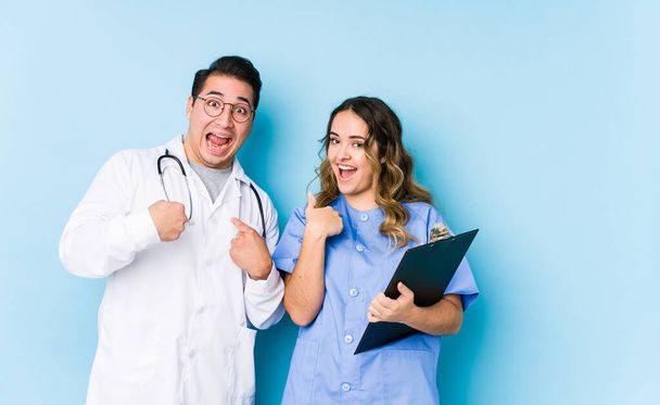 Fiatal orvos pár pózol a kék háttér elszigetelt meglepett mutogatás ujjal, mosolygós széles körben. - Fotó, kép