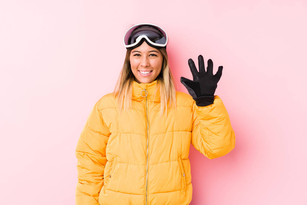 Fiatal kaukázusi nő visel sí ruhák rózsaszín háttér mosolygós vidám mutatja az ötös számú ujjaival. - Fotó, kép