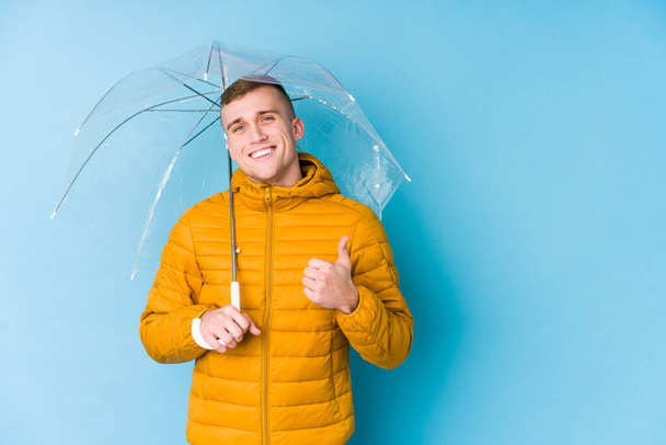 Молодий білий чоловік тримає парасольку посміхаючись і піднімаючи великий палець вгору
 - Фото, зображення