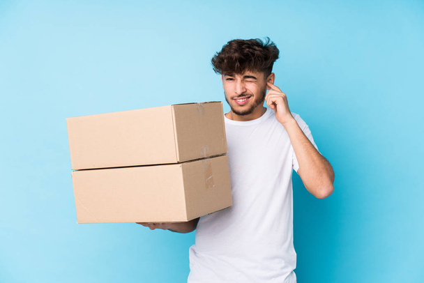 Young arab man holding boxes isolated covering ears with hands. - 写真・画像