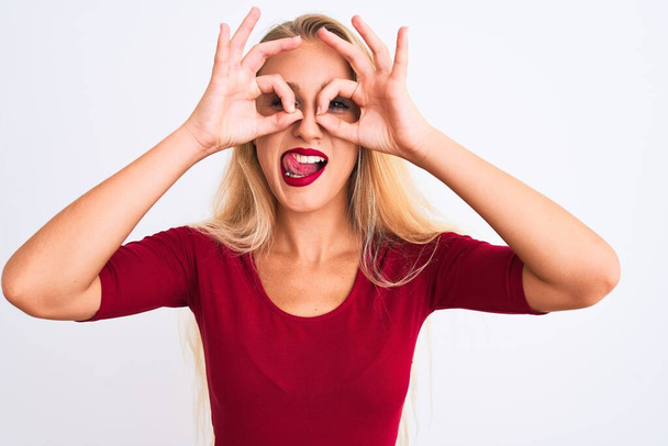 Young beautiful woman wearing red t-shirt standing over isolated white background doing ok gesture like binoculars sticking tongue out, eyes looking through fingers. Crazy expression. - Photo, Image
