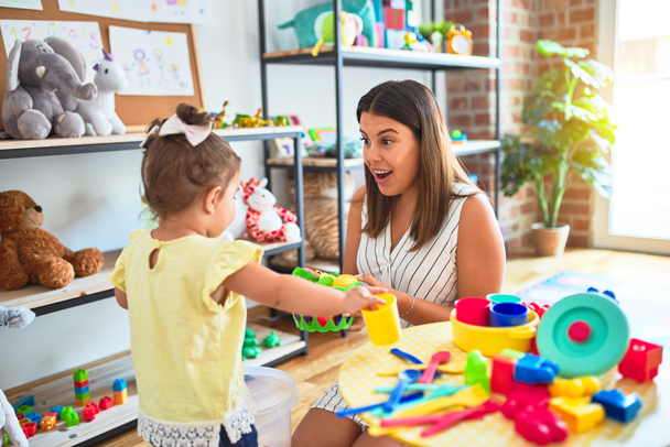 Молодой красивый учитель и ребенок играет с блюдами, столовыми приборами и кубками игрушки на столе в детском саду
 - Фото, изображение