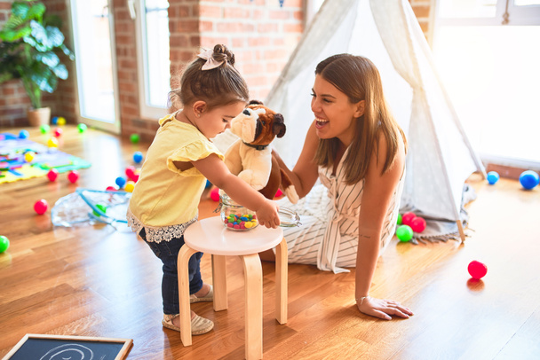 Jovem e bela professora brincando com boneca de cachorro e criança segurando jarro de bolas de chocolate no jardim de infância
 - Foto, Imagem