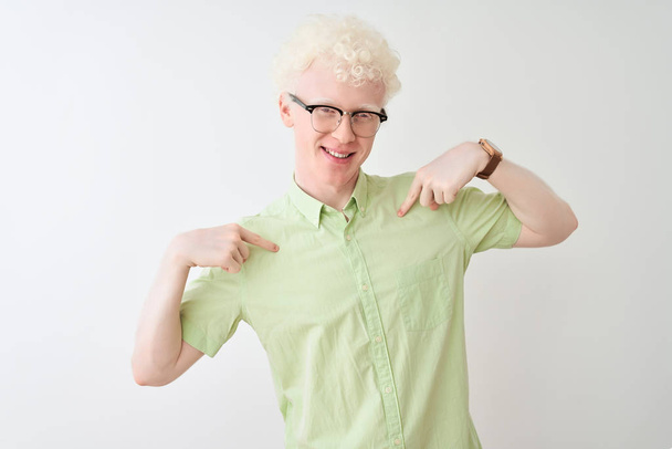 Nuori albiino vaalea mies yllään vihreä paita ja lasit yli eristetty valkoinen tausta näyttää luottavainen hymyillen kasvot, osoittaa itseään sormilla ylpeä ja onnellinen
. - Valokuva, kuva