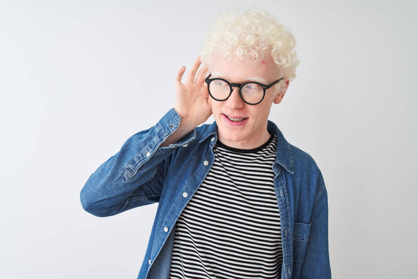 Nuori albiino vaalea mies yllään denim paita ja lasit yli eristetty valkoinen tausta hymyilee käsi korva kuunnellen kuulo huhu tai juoruja. Kuurous-käsite
. - Valokuva, kuva