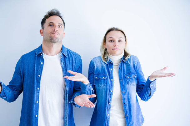 Young beautiful couple wearing denim shirt standing over isolated white background clueless and confused expression with arms and hands raised. Doubt concept. - Photo, Image