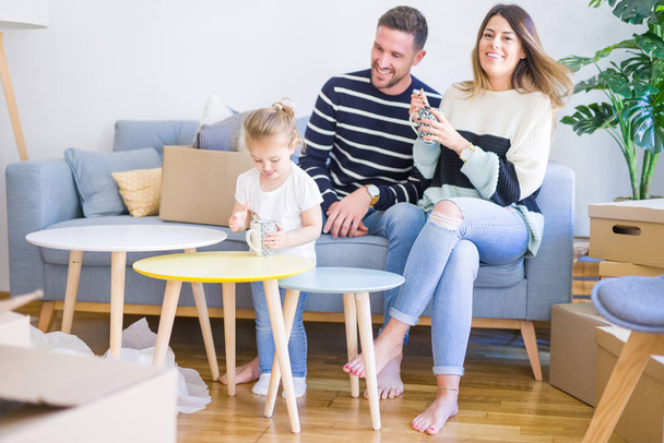 schöne Familie, Eltern sitzen auf dem Sofa und trinken Kaffee und schauen seinem Kind zu, wie es im neuen Zuhause um Pappkartons spielt - Foto, Bild
