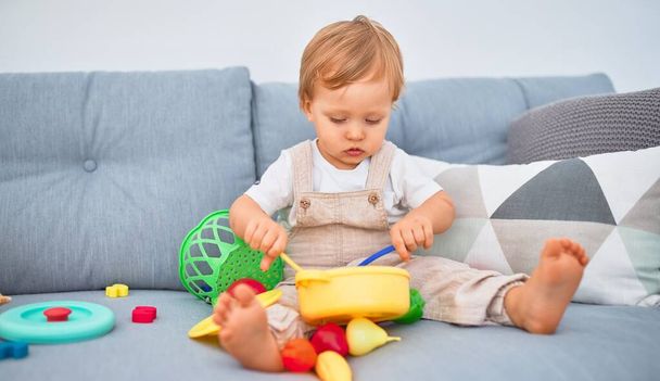 Uroczy blond maluch siedzi na kanapie i bawi się plastikowymi zabawkami w domu - Zdjęcie, obraz