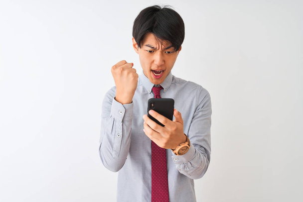 中国のビジネスマンは、孤立した白い背景の上に立ってスマートフォンを使用してネクタイを身に着けている怒りといらいら叫び、狂気と怒りの手で叫んで、怒りの概念 - 写真・画像