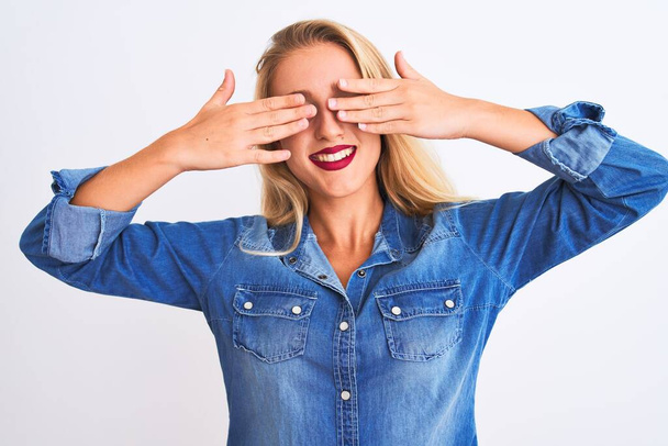 Young beautiful woman wearing casual denim shirt standing over isolated white background covering eyes with hands smiling cheerful and funny. Blind concept. - Photo, Image