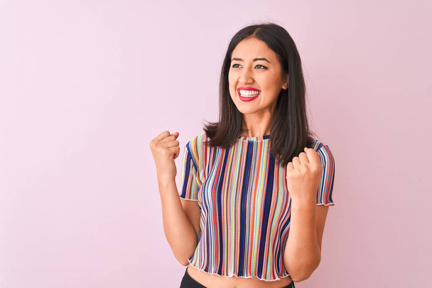 Young chinese woman wearing colorful striped t-shirt standing over isolated pink background very happy and excited doing winner gesture with arms raised, smiling and screaming for success. Celebration concept. - Photo, Image