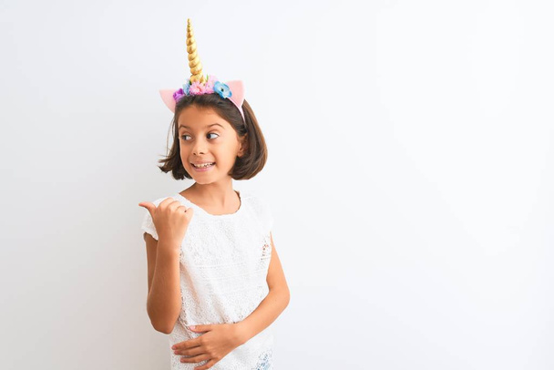 mooi kind meisje dragen eenhoorn diadeem staande over geïsoleerde witte achtergrond glimlachen met gelukkig gezicht kijken en wijzen naar de zijkant met duim omhoog. - Foto, afbeelding