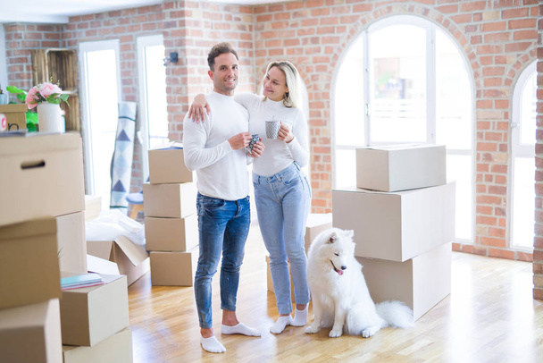 Young beautiful couple with dog standing drinking cup of coffee at new home around cardboard boxes - Photo, Image