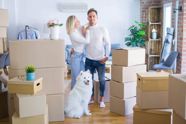 Young beautiful couple with dog standing at new home around cardboard boxes - Photo, Image