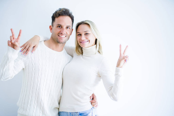 Young beautiful couple wearing casual t-shirt standing over isolated white background smiling with happy face winking at the camera doing victory sign. Number two. - 写真・画像