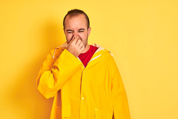 Young man wearing rain coat standing over isolated yellow background smelling something stinky and disgusting, intolerable smell, holding breath with fingers on nose. Bad smells concept. - Photo, Image