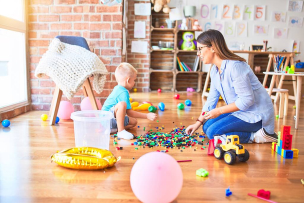 Young caucasian child playing at playschool with teacher. Mother and son at playroom bulding a tower with toy blocks - Foto, Bild