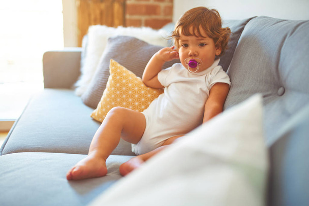 Красива дівчинка-малюк в білій футболці, що сидить на дивані, використовуючи соску
 - Фото, зображення