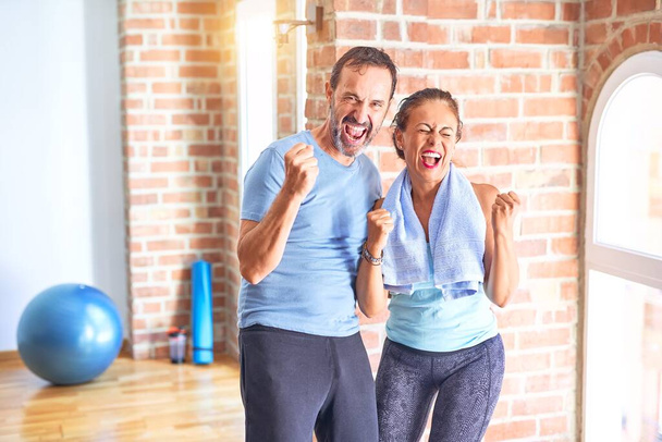 Couple sportif d'âge moyen debout avant de faire de l'exercice à la salle de gym très heureux et excité de faire geste gagnant avec les bras levés, souriant et criant pour le succès. Concept de célébration
. - Photo, image
