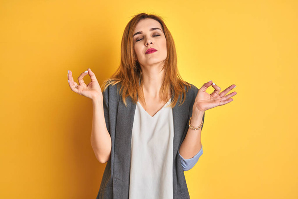 Redhead caucasian business woman over yellow isolated background relaxed and smiling with eyes closed doing meditation gesture with fingers. Yoga concept. - Photo, Image