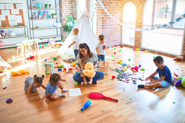 Όμορφη δασκάλα και ομάδα μικρών παιδιών που παίζουν γύρω από πολλά παιχνίδια στο νηπιαγωγείο - Φωτογραφία, εικόνα