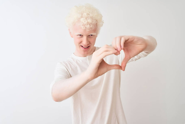 junger Albino-blonder Mann in lässigem T-Shirt, der vor isoliertem weißem Hintergrund steht und verliebt lächelt und Herzsymbolform mit den Händen macht. Romantisches Konzept. - Foto, Bild