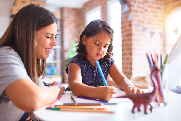 Güzel öğretmen ve küçük kız anaokulunda renkli kalemlerle resim çiziyor. - Fotoğraf, Görsel