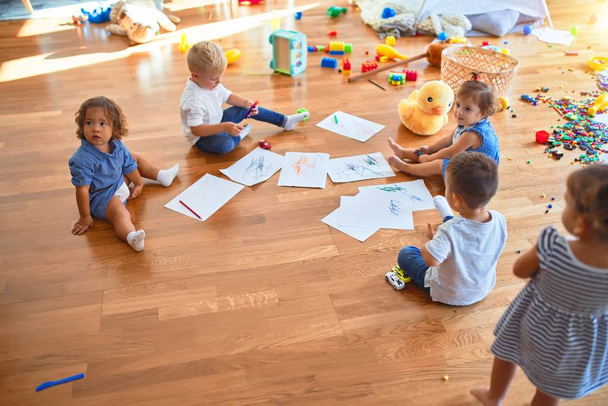 Αξιολάτρευτη ομάδα μικρών παιδιών που κάθονται στο πάτωμα ζωγραφίζοντας χρησιμοποιώντας χαρτί και μολύβι γύρω από πολλά παιχνίδια στο νηπιαγωγείο - Φωτογραφία, εικόνα