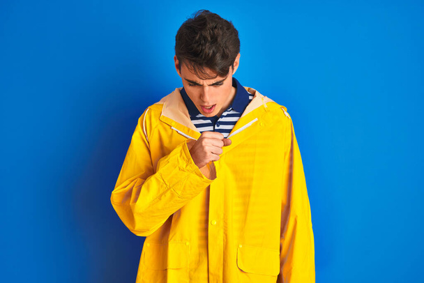 Έφηβος ψαράς αγόρι φορώντας κίτρινο αδιάβροχο πάνω από απομονωμένο υπόβαθρο αίσθημα αδιαθεσίας και βήχα ως σύμπτωμα για το κρύο ή βρογχίτιδα. Έννοια υγειονομικής περίθαλψης. - Φωτογραφία, εικόνα