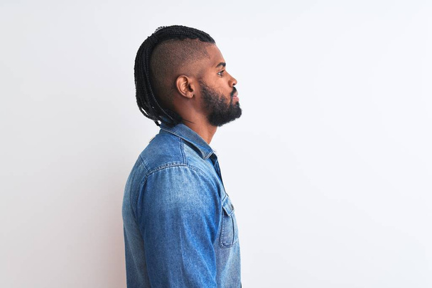 Afrikanisch-amerikanischer Mann mit Zöpfen trägt Jeanshemd über isoliertem weißem Hintergrund zur Seite schauen, entspannen Profil Pose mit natürlichem Gesicht mit selbstbewusstem Lächeln. - Foto, Bild