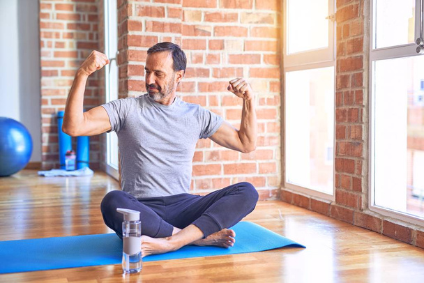 Middelbare leeftijd knappe sportman zittend op mat doen stretching yoga oefening op sportschool tonen armen spieren glimlachend trots. Fitness concept. - Foto, afbeelding