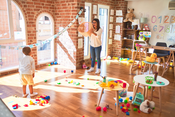 Beautiful teacher and toddler playing basketball using ball and wicker basket around lots of toys at kindergarten - Photo, Image