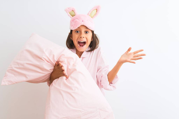 Child girl wearing sleep mask and pajama holding pillow over isolated white background very happy and excited, winner expression celebrating victory screaming with big smile and raised hands - 写真・画像