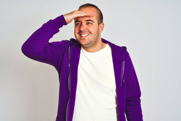 孤立した白い背景の上に立って紫色のスウェットシャツを着て若い男は非常に幸せと笑みを浮かべて遠くに手を頭の上に見て。検索の概念. - 写真・画像