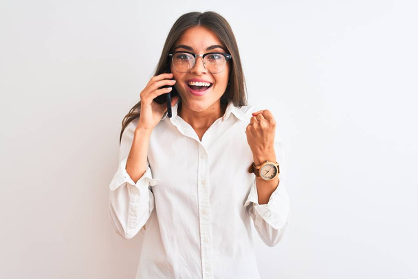 Young businesswoman wearing glasses talking on smartphone over isolated white background screaming proud and celebrating victory and success very excited, cheering emotion - Foto, Imagem