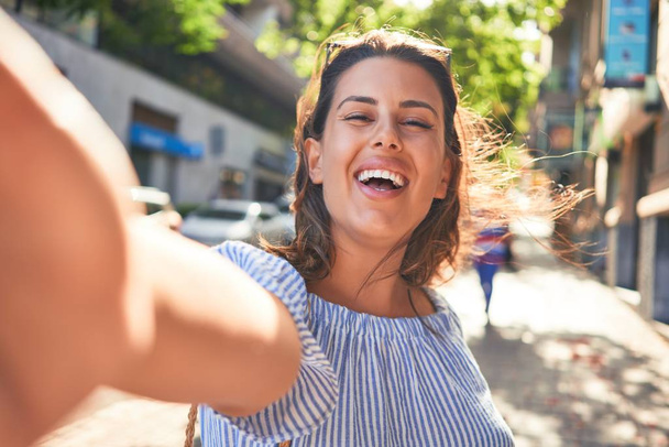 Joven hermosa mujer sonriendo feliz caminando por las calles de la ciudad en un día soleado de verano tomando una foto selfie usando un teléfono inteligente
 - Foto, imagen