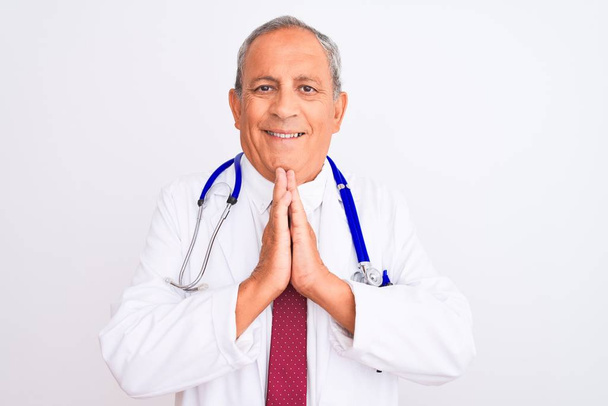 Старший сіро-волохатий лікар у стетоскопі стоїть над ізольованим білим тлом, молячись руками, просячи прощення посміхаючись впевнено
. - Фото, зображення
