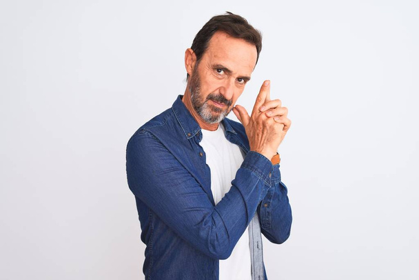Middle age handsome man wearing blue denim shirt standing over isolated white background Holding symbolic gun with hand gesture, playing killing shooting weapons, angry face - Photo, Image