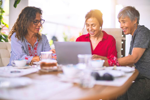 Treffen von Frauen mittleren Alters beim Mittagessen und Kaffeetrinken. Ältere Freunde lächeln glücklich mit Laptop zu Hause an einem sonnigen Tag - Foto, Bild
