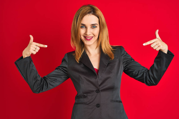 Jonge blanke zakenvrouw draagt een pak over geïsoleerde rode achtergrond en ziet er zelfverzekerd uit met een glimlach op het gezicht, wijzend met de vingers trots en gelukkig. - Foto, afbeelding