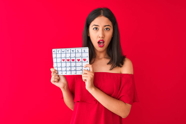 junge schöne Chinesin mit Zeitrahmen-Kalender vor isoliertem rotem Hintergrund erschrocken mit einem Überraschungsgesicht, ängstlich und aufgeregt mit Angstausdruck - Foto, Bild