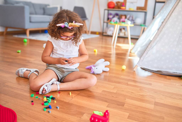 Bela criança usando óculos e diadema de unicórnio sentado no chão brincando com blocos de construção no jardim de infância
 - Foto, Imagem